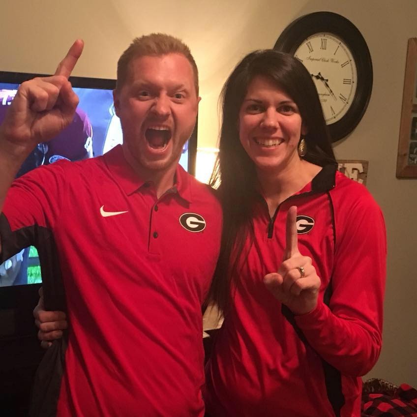 Jason Hughes and his wife Laura Palmer Hughes in matching red shirts with the Georgia Bulldogs logo, both pointing one finger up.